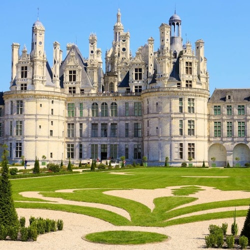 Un grand château avec des tours ornées et un jardin soigné présentant des pelouses et des allées conçues géométriquement.