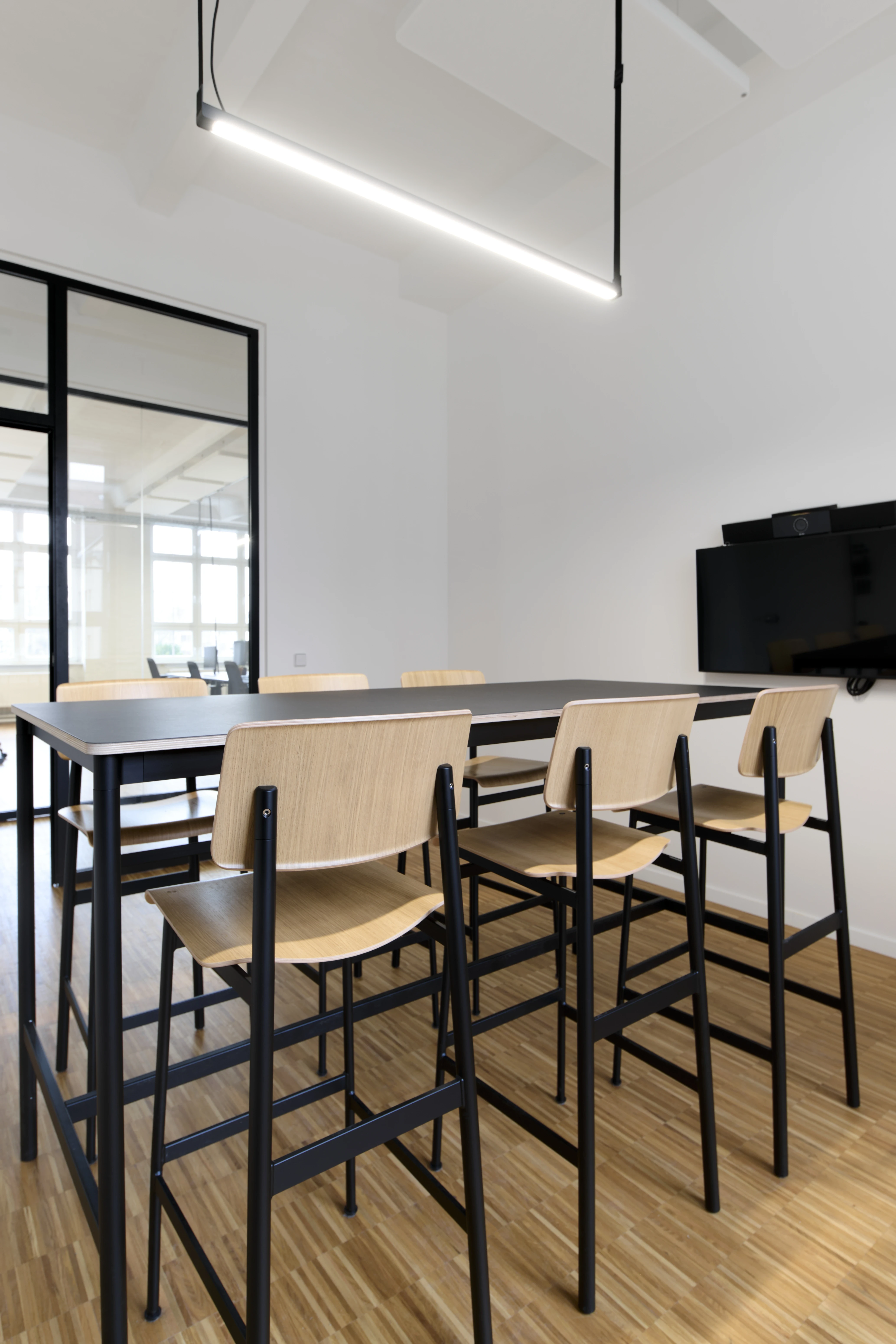 Standing meeting room at PLAN A with a black high table, oak-and-black bar chairs, a wall-mounted TV, a slim linear pendant light and a black-framed glass partition.
