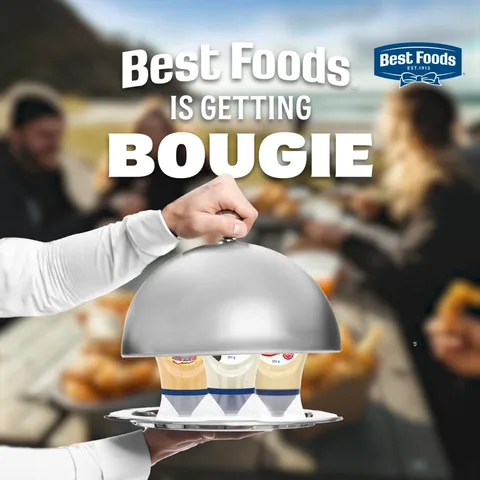 A waiter lifts a silver lid at a picnic, under the words “Best Foods is getting bougie”