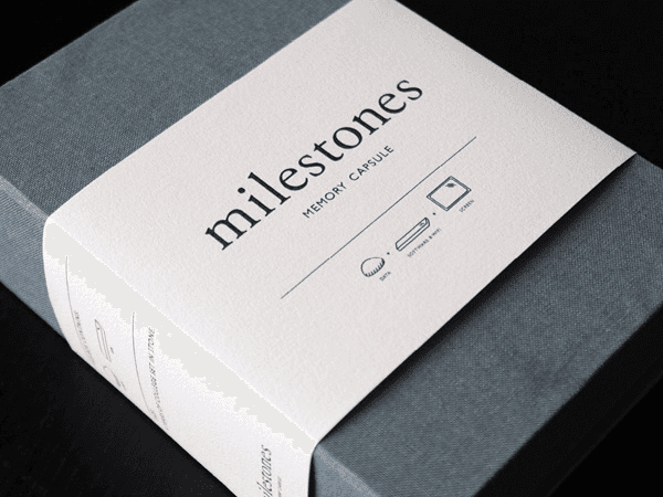 A gray box with a white label displays the brand name "milestones" in a bold, sans-serif font. Below the brand name, the label reads "MEMORY CAPSULE" and includes icons of a soft-sided envelope, a smartphone, and a ball.