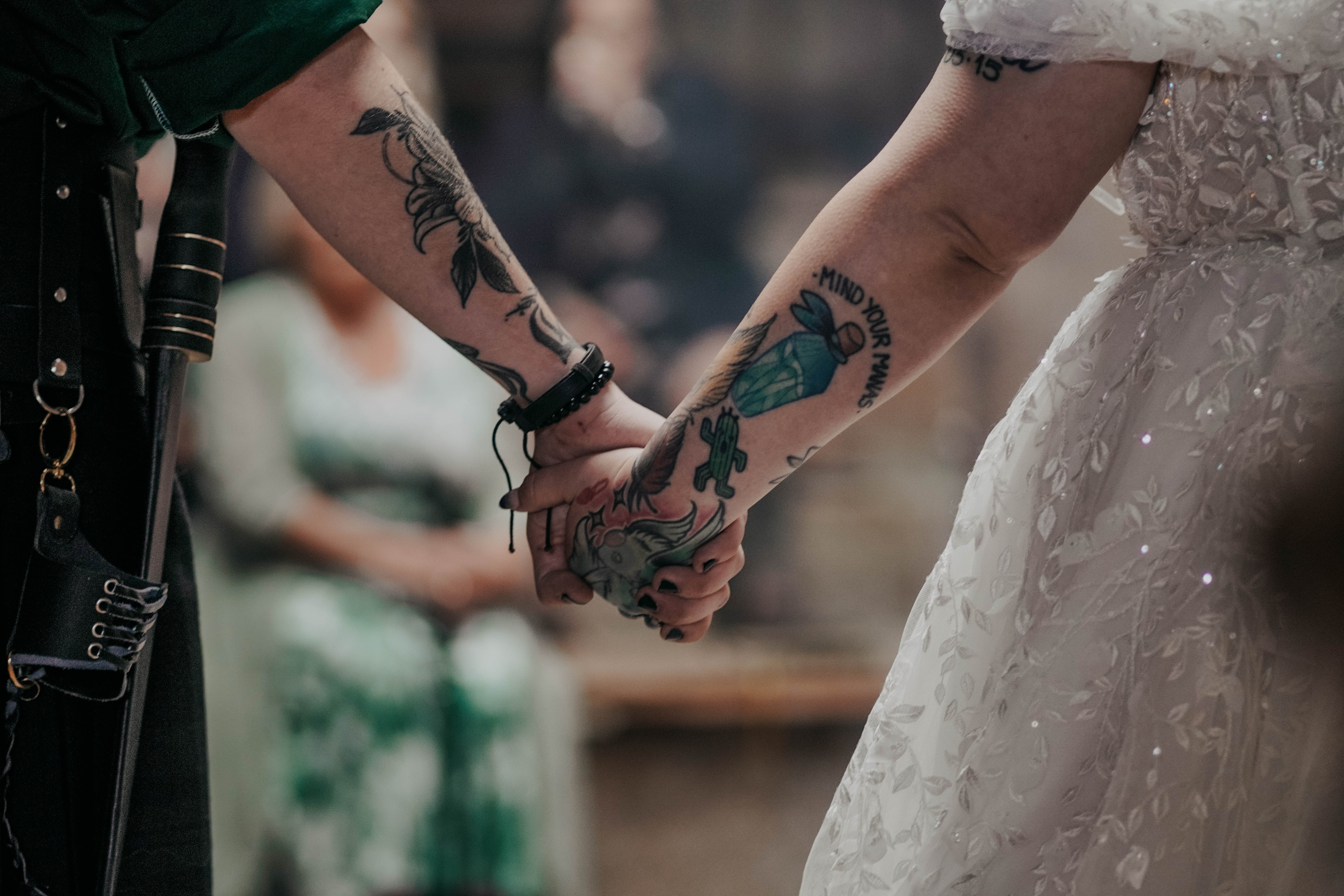 A close-up on the tattooed arms of a bride and groom as they hold hands. The bride wears a white embroidered gown, and the groom is in a dark green shirt. Their clasped hands are the central focus of the image.
