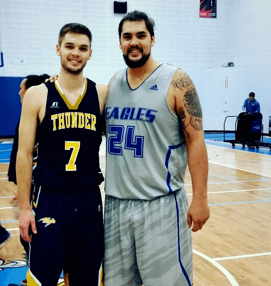 A youth basketball player in an orange jersey dribbling aggressively past a defender during a game, with a referee and spectators visible in the background on an indoor court.