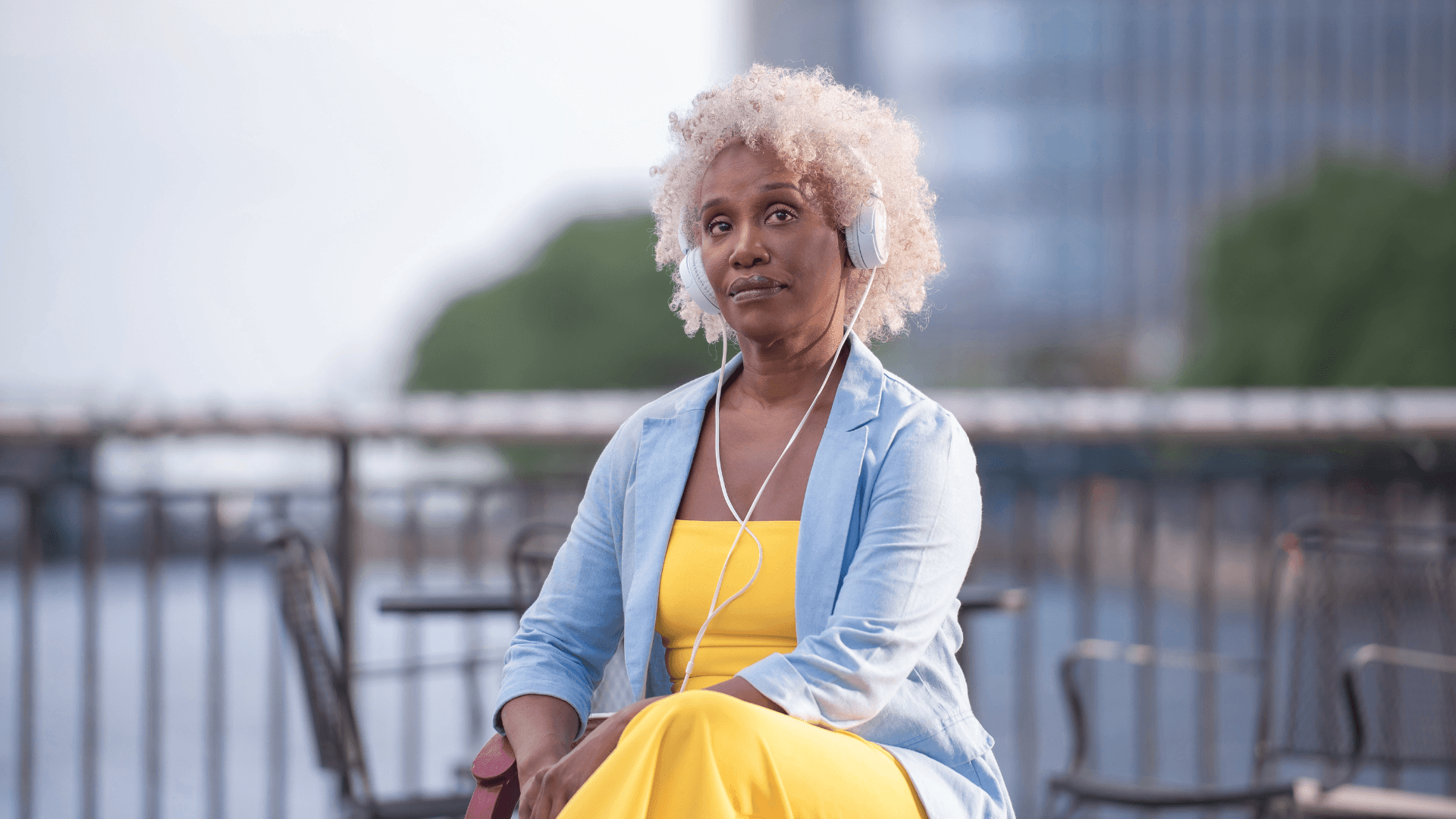 A woman with short, curly hair wears a yellow dress and denim jacket, sitting outdoors near a railing.