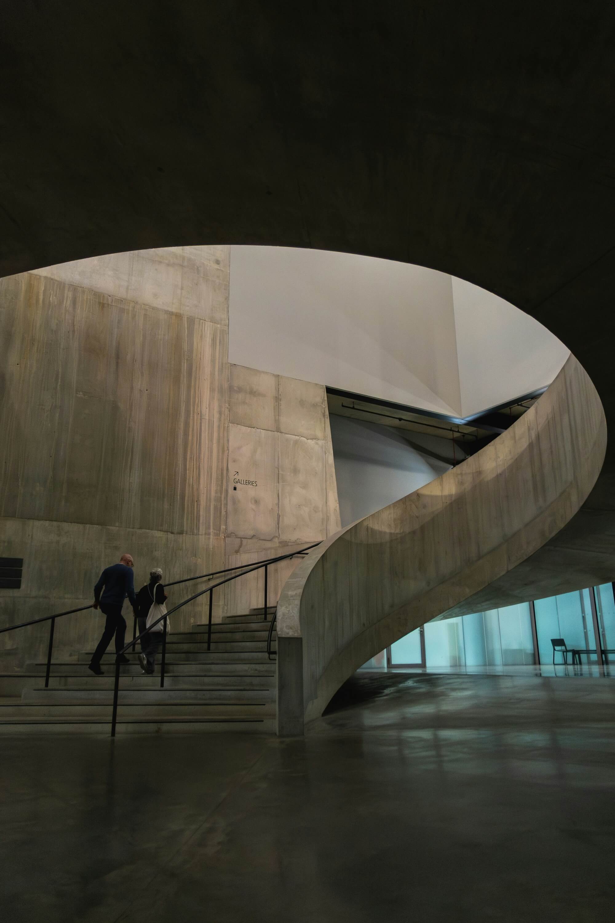 A modern, minimalist interior with a large, curved concrete staircase ascending to the right. Two people walk up, imparting a sense of scale and movement.