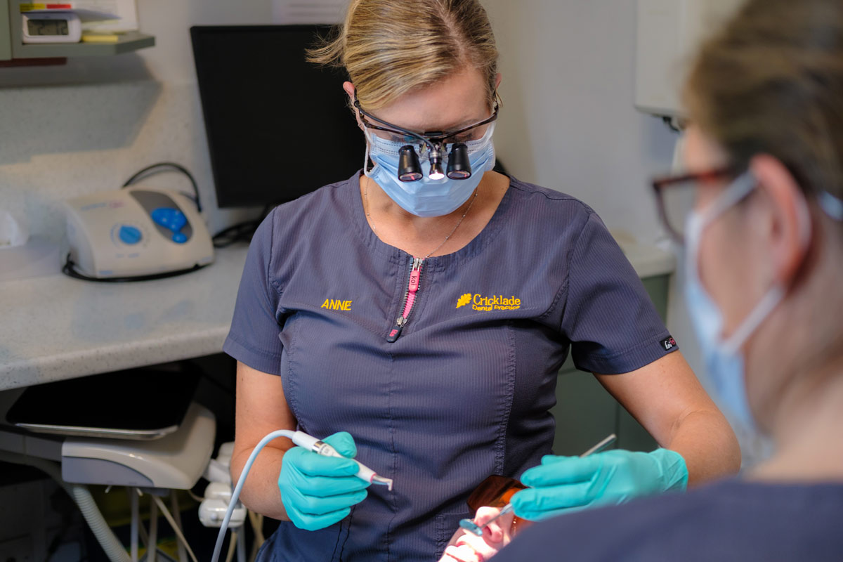A candid shot of Anne, a Dental Hygienist at Cricklade Dental Practice, wearing her dark grey scrub top with her name and the practice logo embroidered. She is wearing a surgical mask, protective eyewear with loupes and a light, and turquoise gloves while treating a patient in a dental chair.