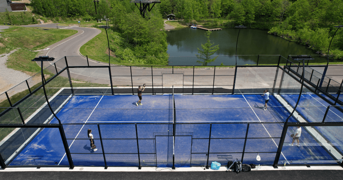 Outdoor blue padel court with players, surrounded by greenery and a lake.