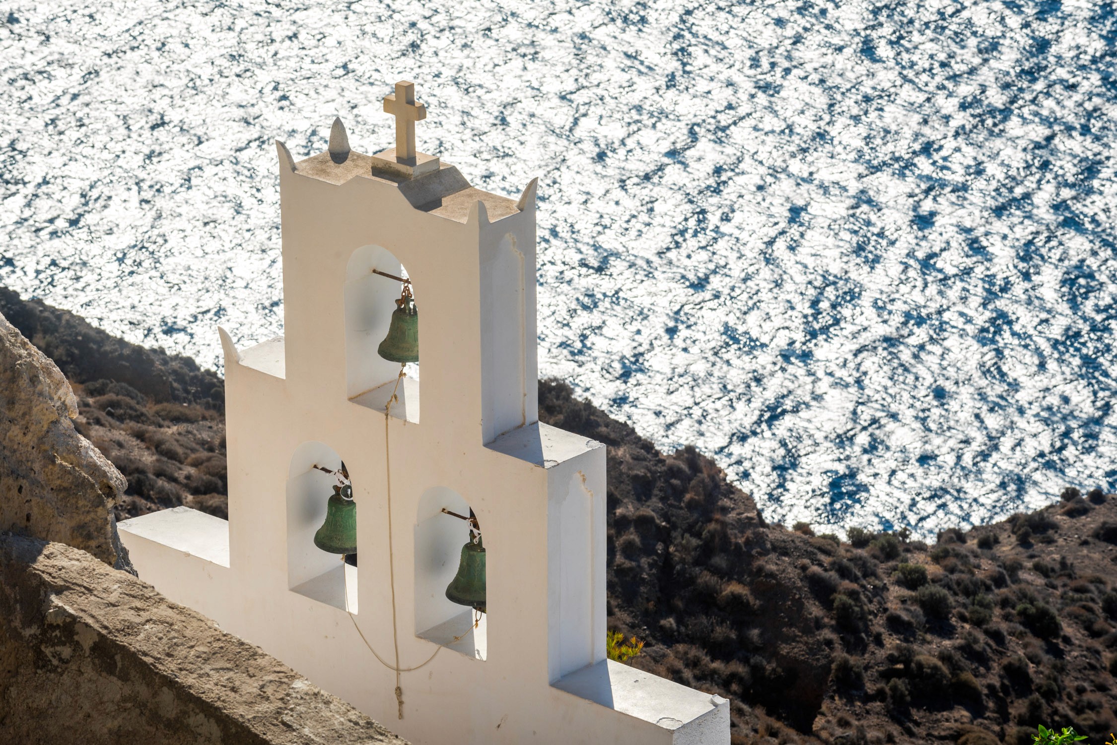 A white stone bell tower with three arched openings and green bells overlooks a blue sea with white waves.