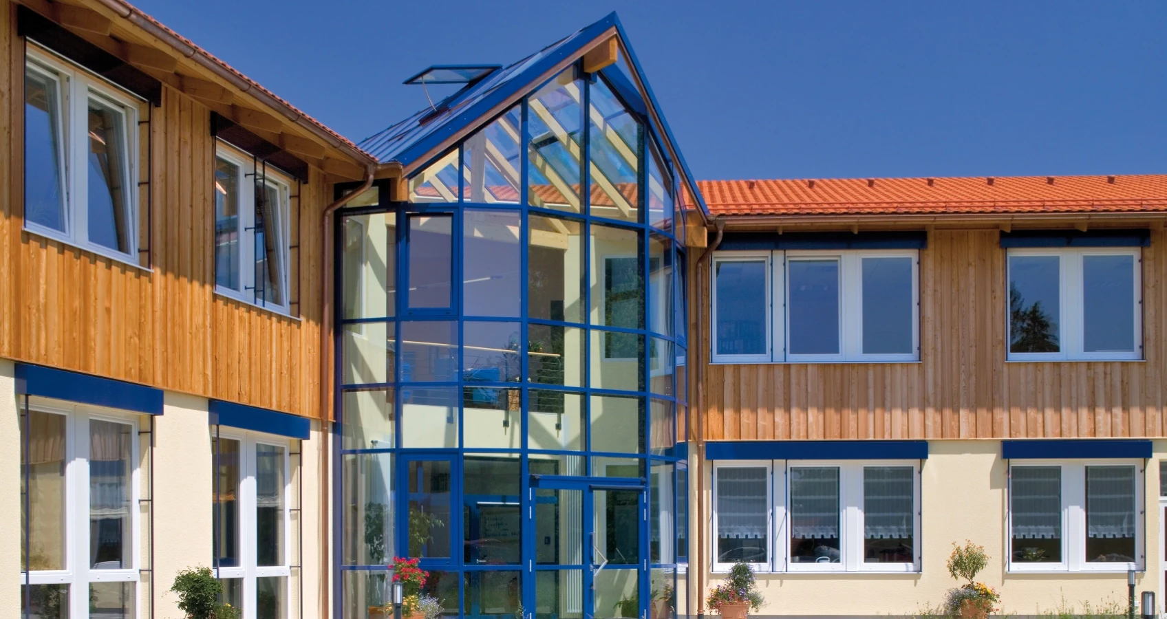Modernes Gebäude mit Holzfassade, Glasgiebel und vielen Fenstern vor strahlend blauem Himmel.