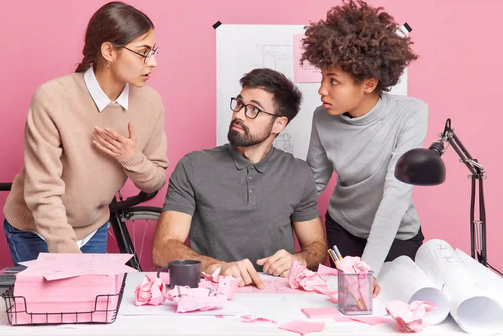 A frustrated creative team experiences a project setback while collaborating at a desk covered in crumpled pink paper.