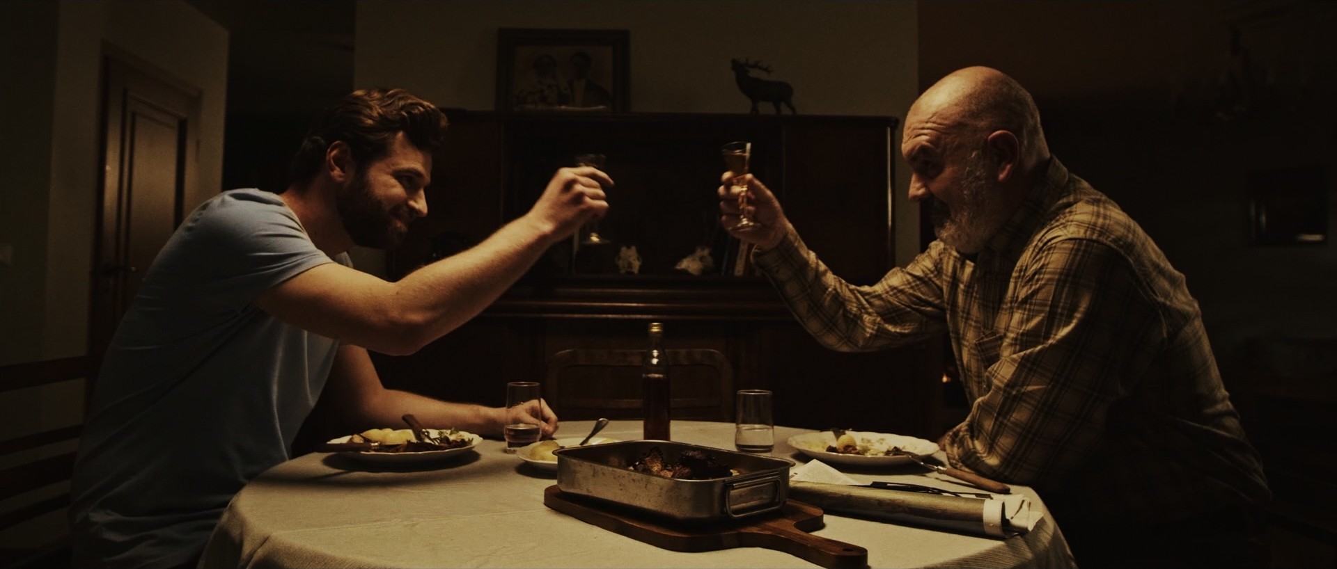 Cinematic interior shot of two men drinking at a table, low-key lighting.