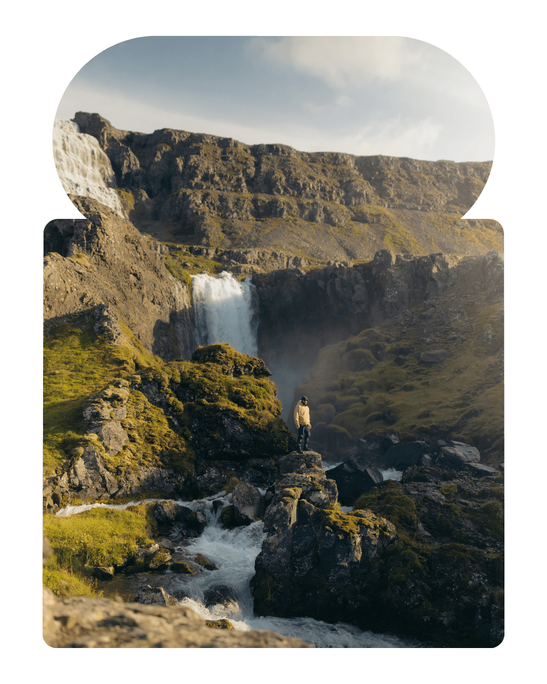 Winding road through green hills with parked cars and small buildings beside a river in a wide rural landscape.