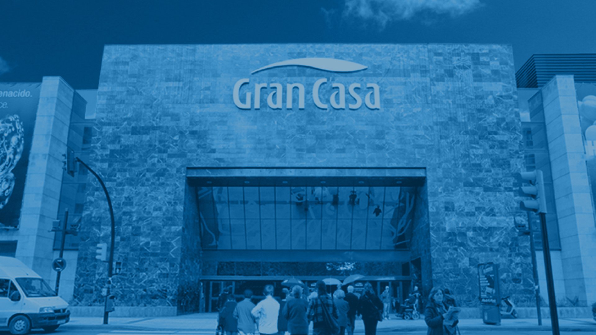 Facade of a large shopping mall with "Gran Casa" sign. Marble exterior, glass entrance. People walk towards it. Blue-tinted image, clear sky.