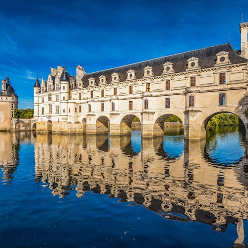 Un pont en pierre avec plusieurs arches s'étend au-dessus d'une rivière calme, reflétant un grand château orné sous un ciel bleu éclatant.