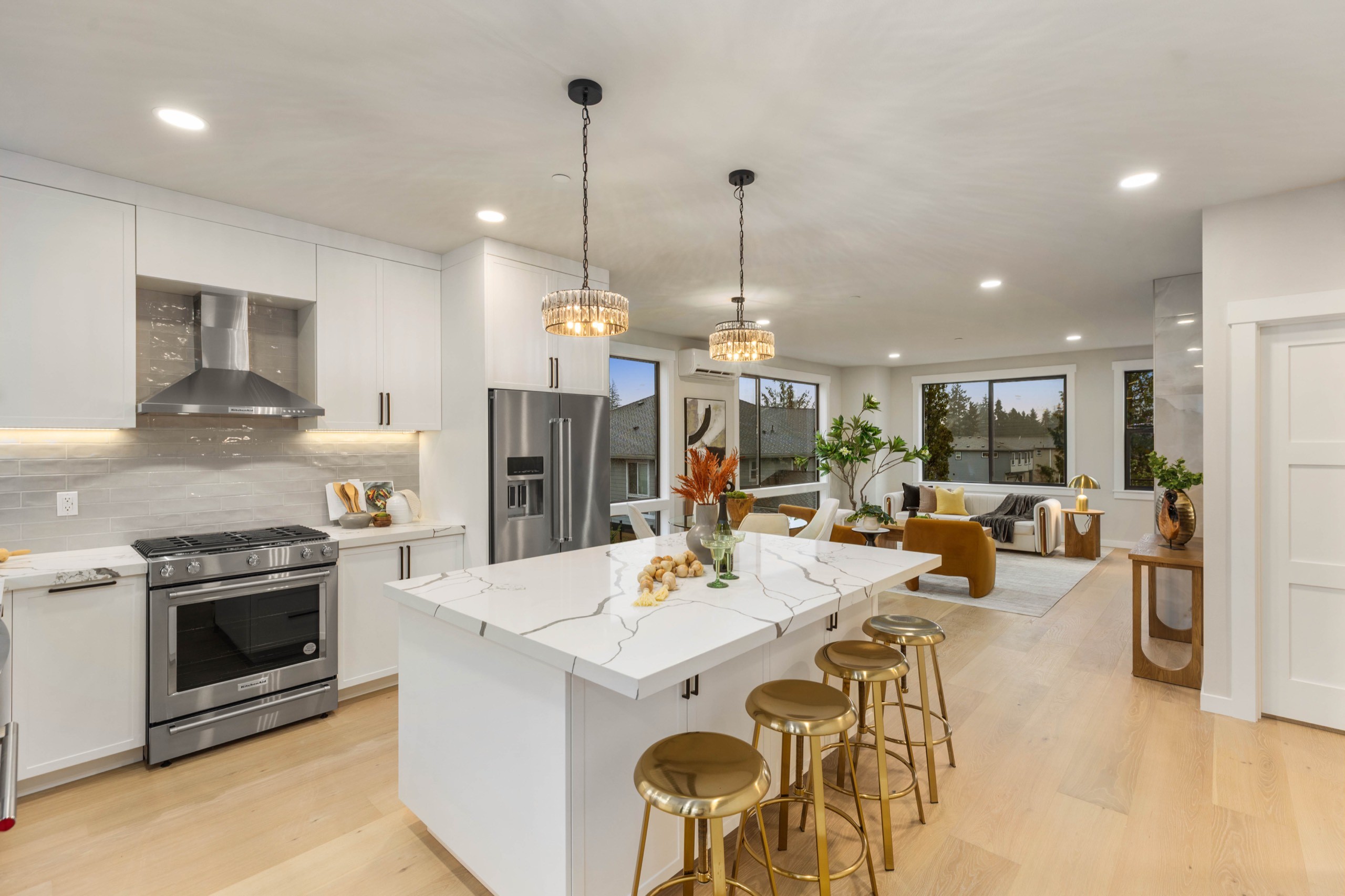 Modern open-concept luxury kitchen with large white island, gold bar stools, stainless steel appliances, pendant lighting, and seamless flow into living area