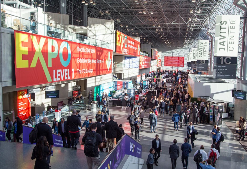 Crowds walk through a large convention center expo hall with banners, exhibits, and booths visible.