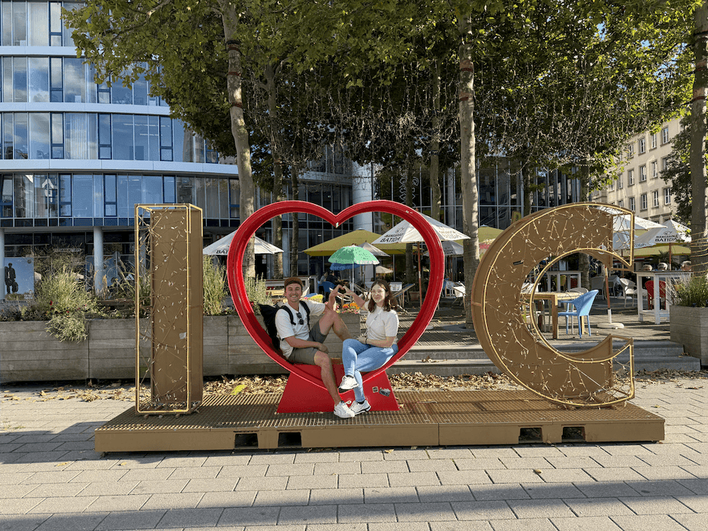 Große Skulptur mit den Buchstaben I und C, dazwischen ein rotes Herz, auf dem zwei Personen sitzen, im Hintergrund moderne Gebäude und Bäume