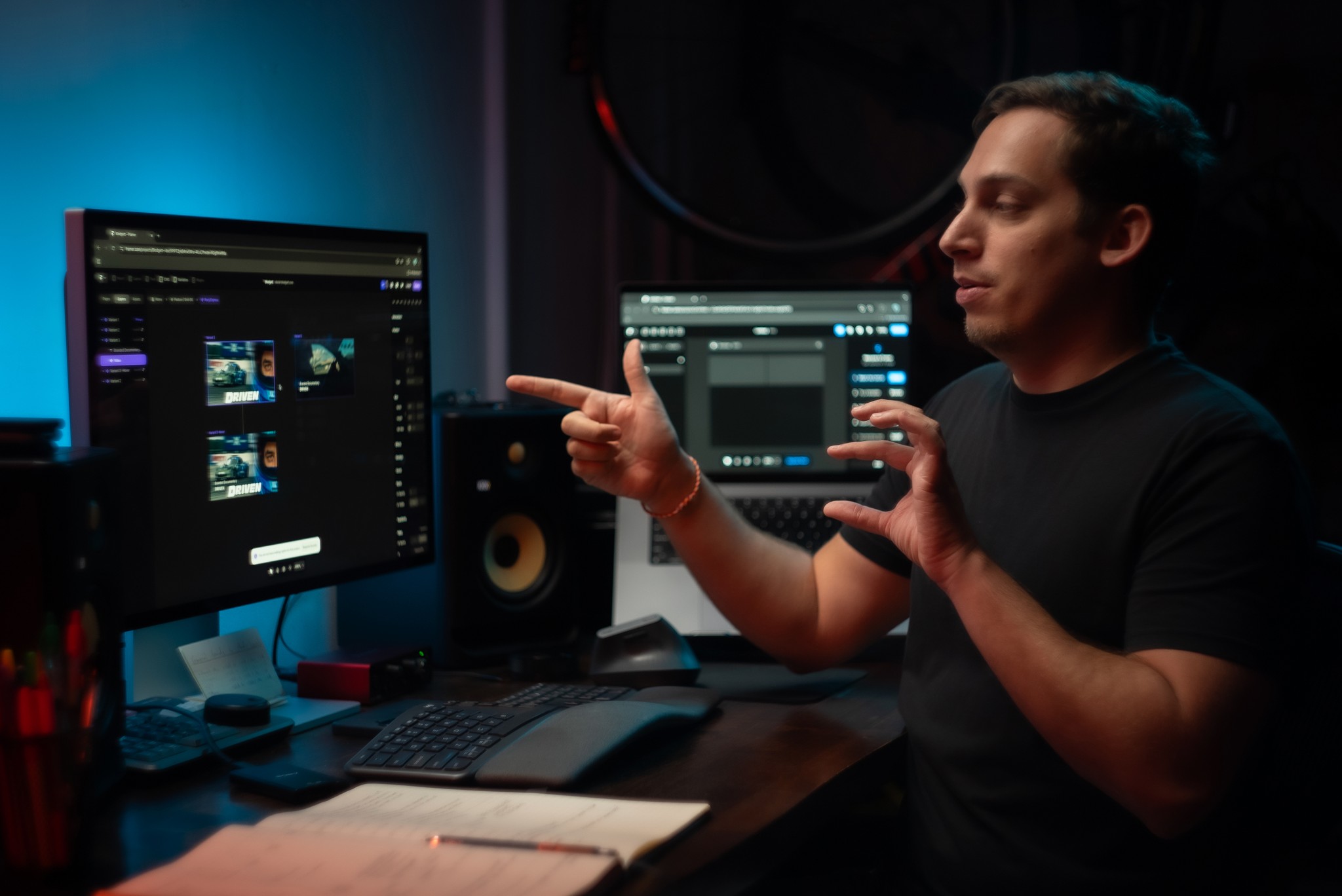 A web designer in a black t-shirt gestures with his hands while explaining Framer techniques at his desk. He sits in a dimly lit room with blue ambient lighting, facing dual monitors displaying dark-mode interface designs.