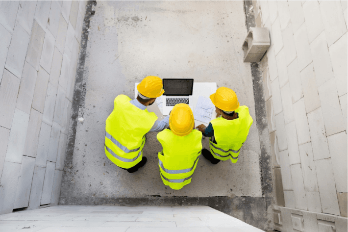 A top-down view of three construction workers on-site using a laptop to look at data