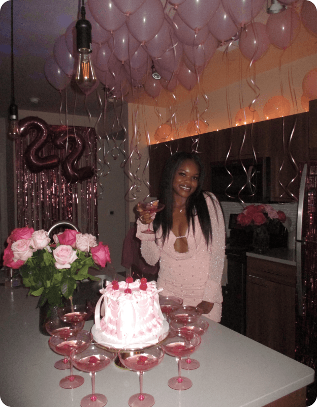 A woman celebrates at a decorated table with a cake and flowers, surrounded by pink balloons and festive decor.