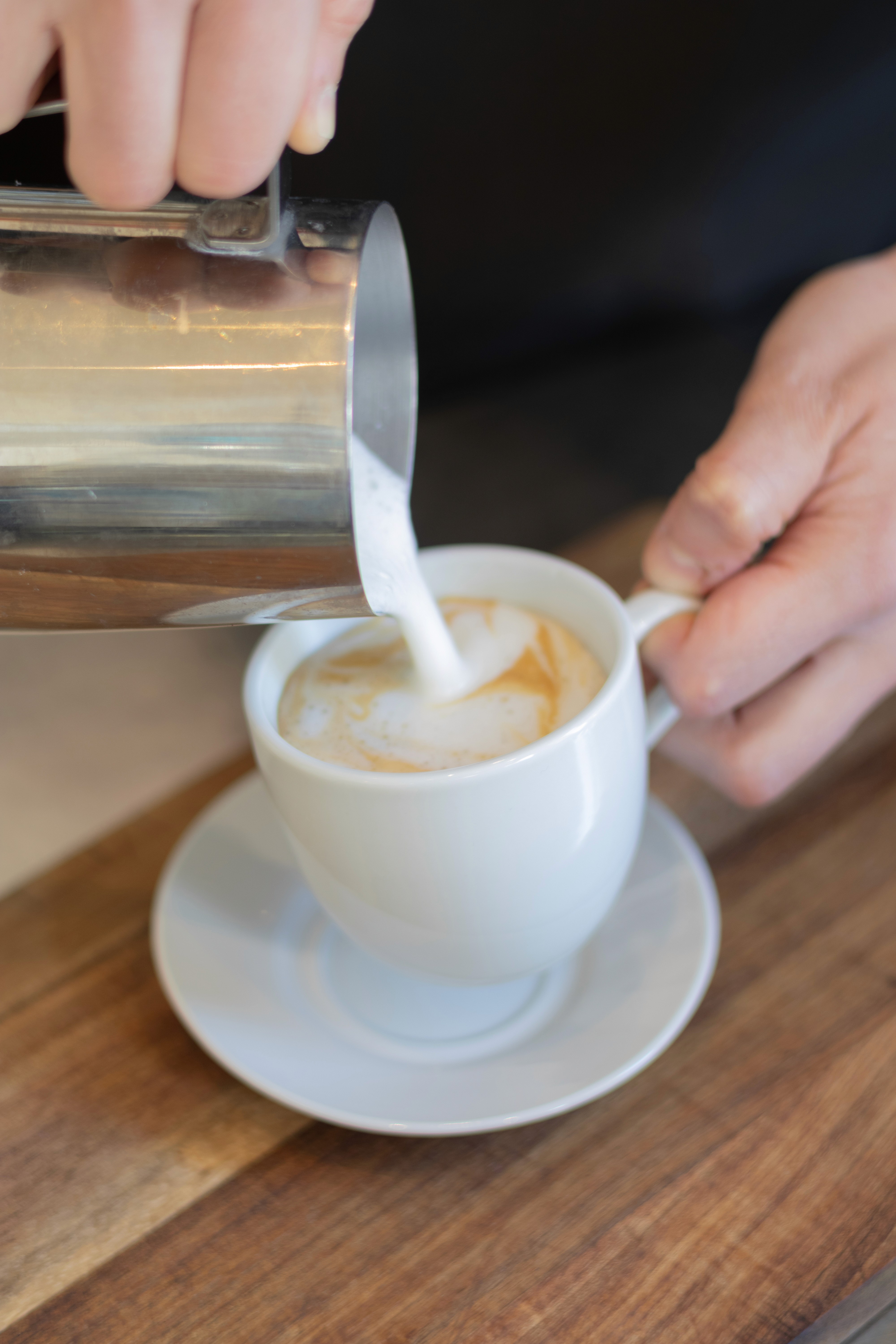 Zubereitung eines Capuccinos, eine Servicekraft hält die Tasse sowie das Milchkännchen in der Hand und lässt die MIlch hineingleiten