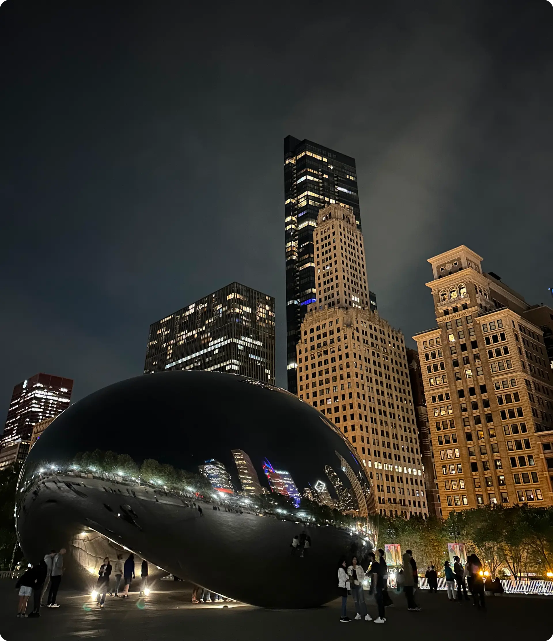 A nighttime view of the Cloud Gate sculpture, also known as "The Bean," in Chicago's Millennium Park. The large, reflective, bean-shaped structure mirrors the surrounding city skyline, including tall buildings with illuminated windows. People are gathered around the sculpture, taking photos and enjoying the scene.