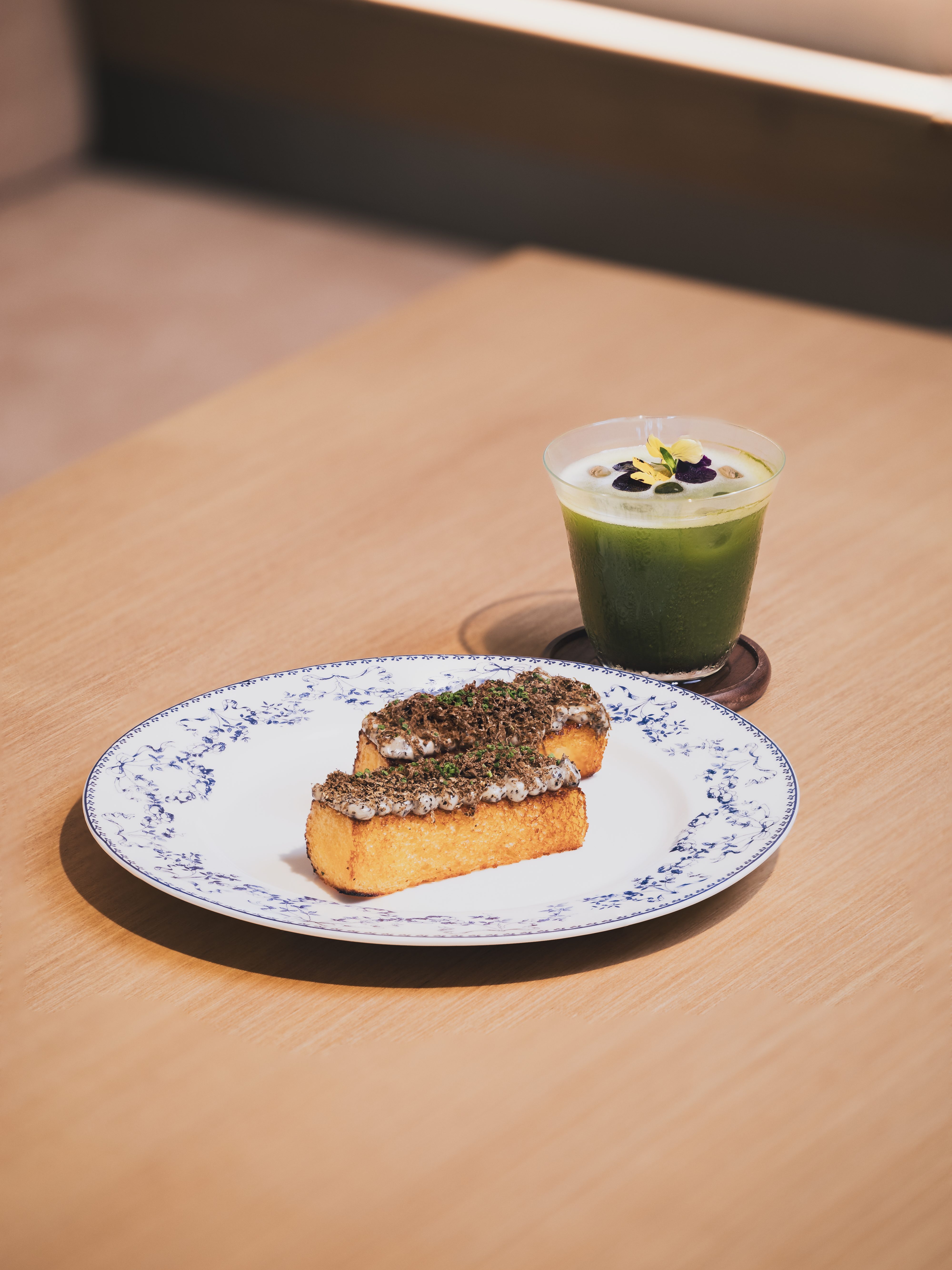 A plate of toasted bread topped with a rich spread, accompanied by a green cocktail garnished with edible flowers, set on a wooden table.