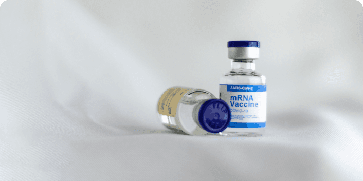 Two small glass medical vials on a white fabric surface, with the front vial labelled "mRNA Vaccine – SARS-CoV-2 – COVID-19" with a blue and silver cap.