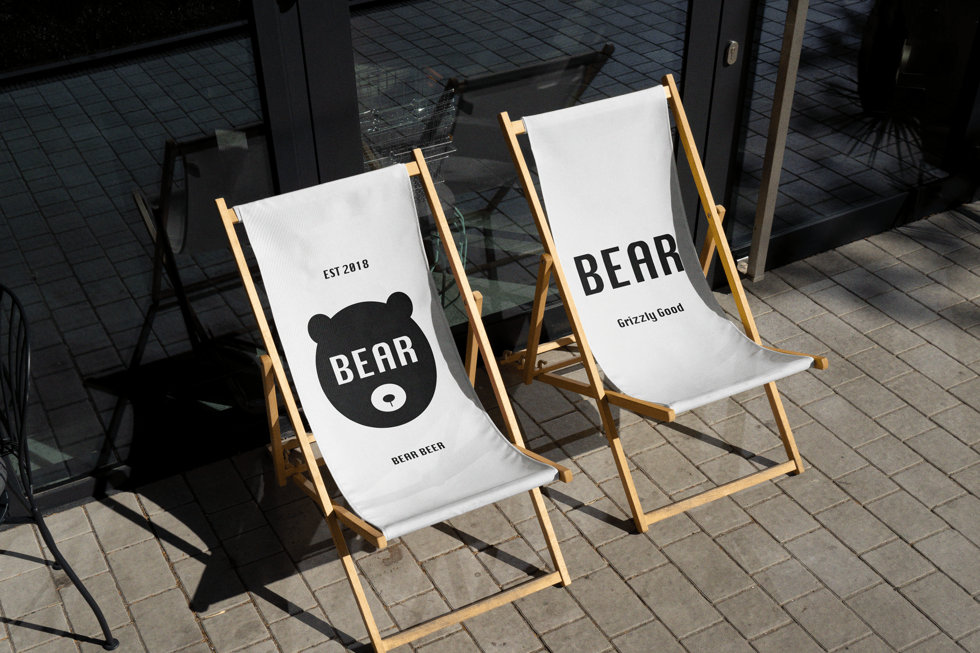 Two wooden lounge chairs with white canvas seats and the 'BEAR' logo printed in large letters on one of the seats, with a small bear logo below it. The chairs are placed outside a cafe or brewery, on a stone patio.