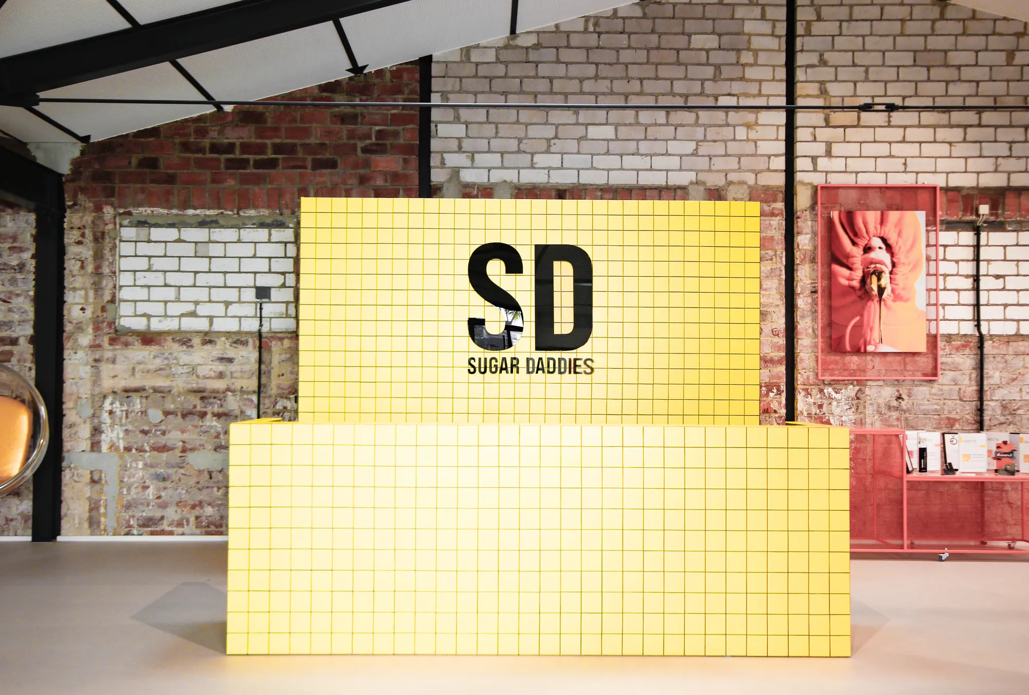 Yellow tiled reception counter with SD logo in front of a brick wall and bubble chair.