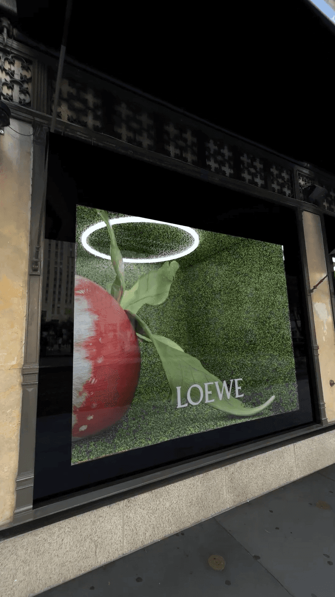 A digital Loewe window display showing a large fruit and a glowing halo against a green wall