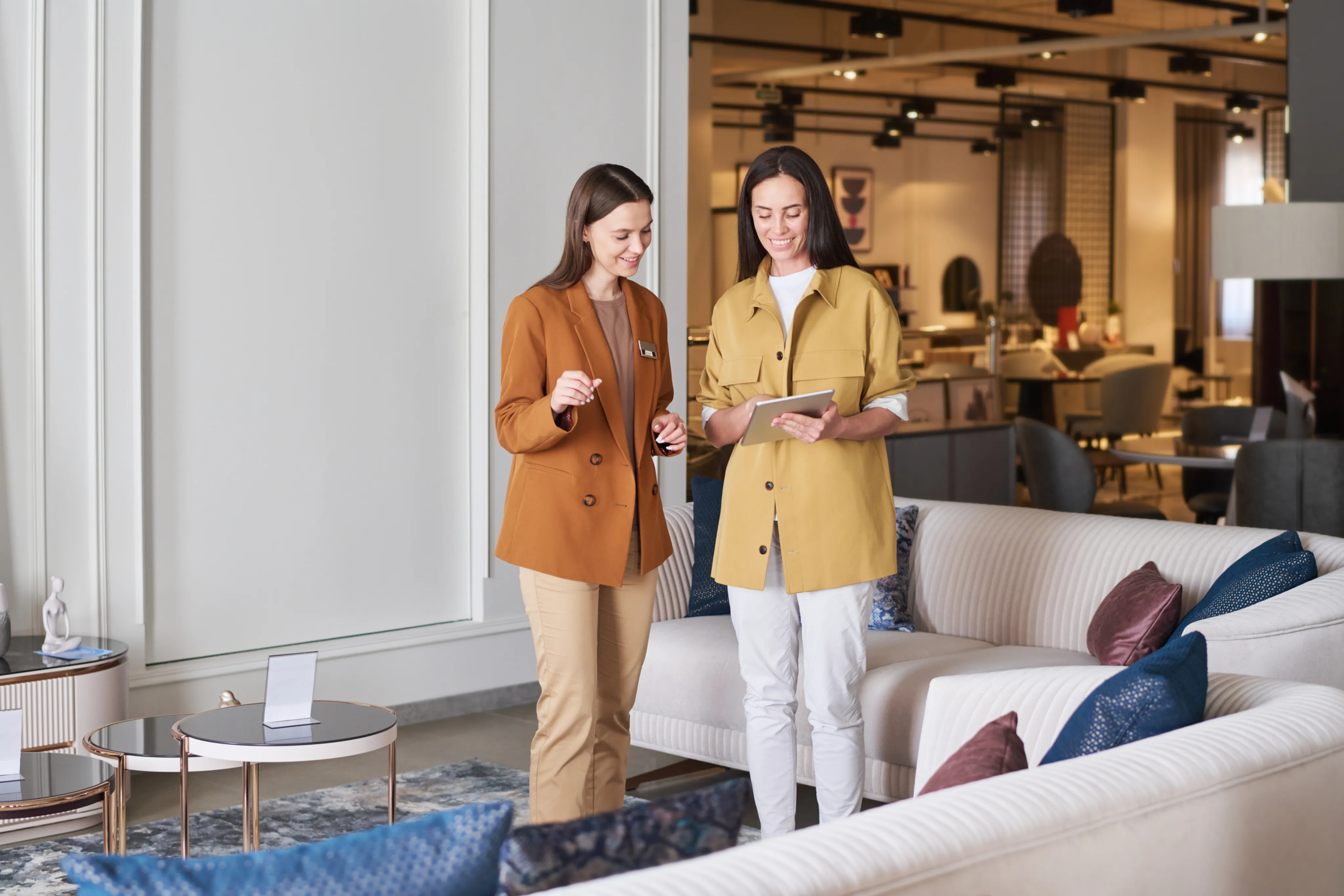 Two women in casual jackets stand in a well-lit furniture showroom, discussing with a tablet in hand. Sofas and decor pieces surround them, creating a professional and modern atmosphere.