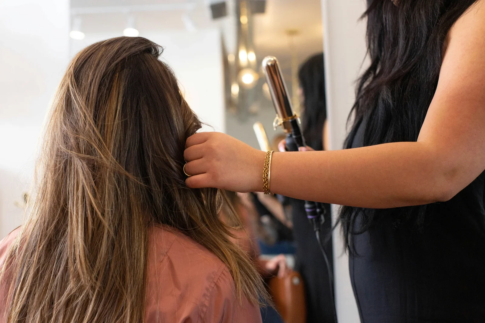 Hairstylist curling client's hair with curling iron in salon mirror setting.