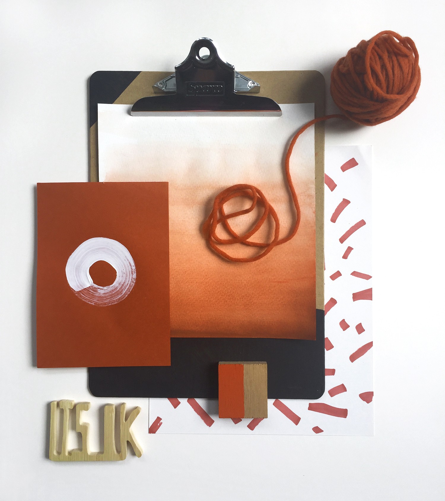 Assemblage of objects: clipboard with ombre rust rest paint, yarn, painted block, and rust red paper with handpainted white circle, and "It's Ok" wood carving