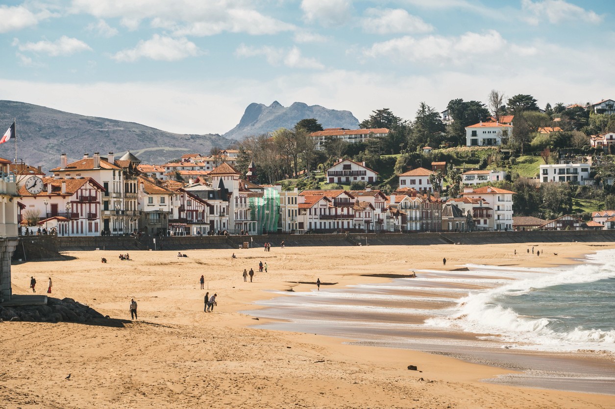 Saint Jean de Luz beachfront view of a town with colorful buildings, mountains in the background, and people enjoying the beach.