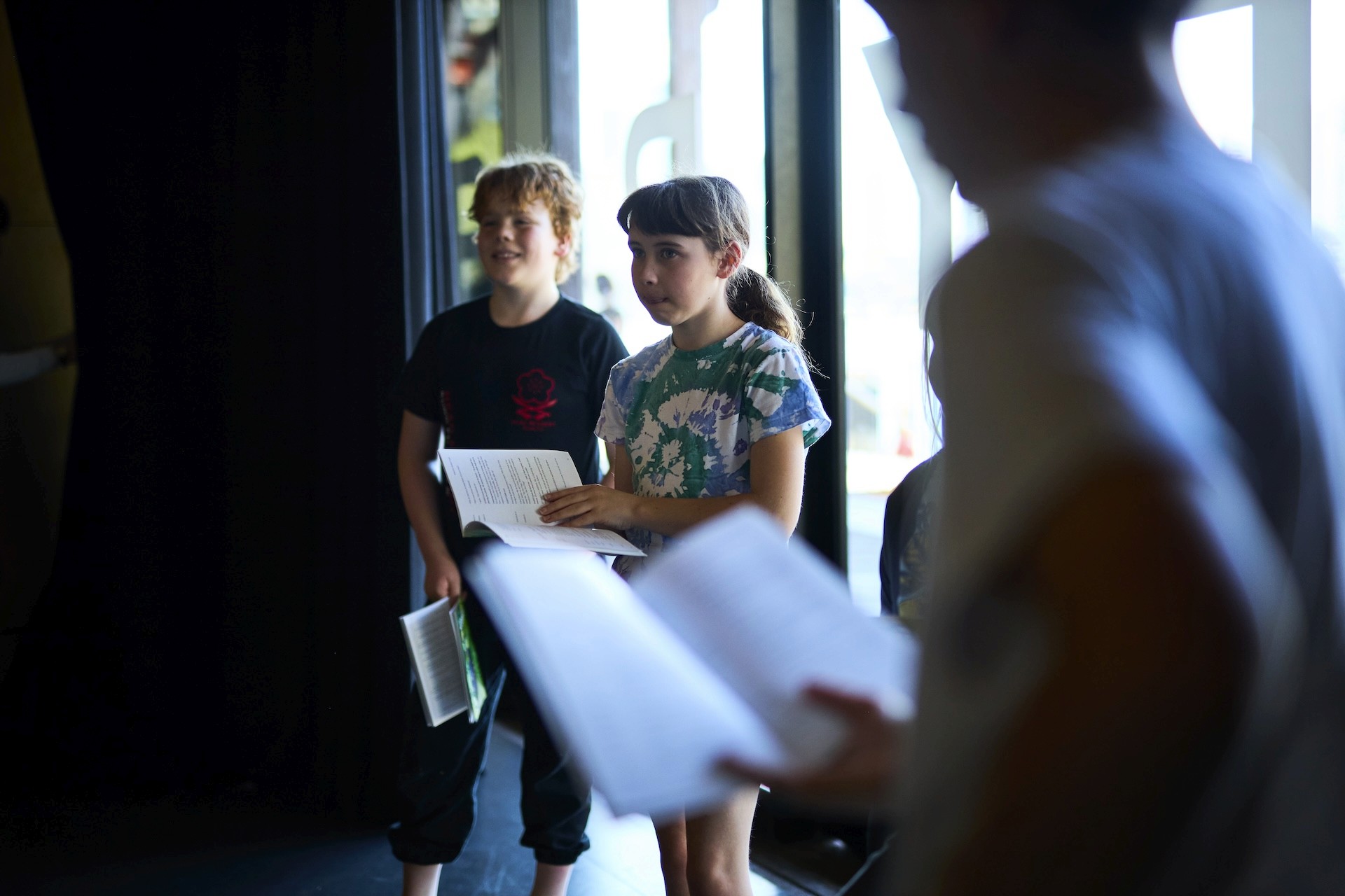 Two children stand near a window holding scripts while listening during a reading exercise, with other participants blurred in the foreground.
