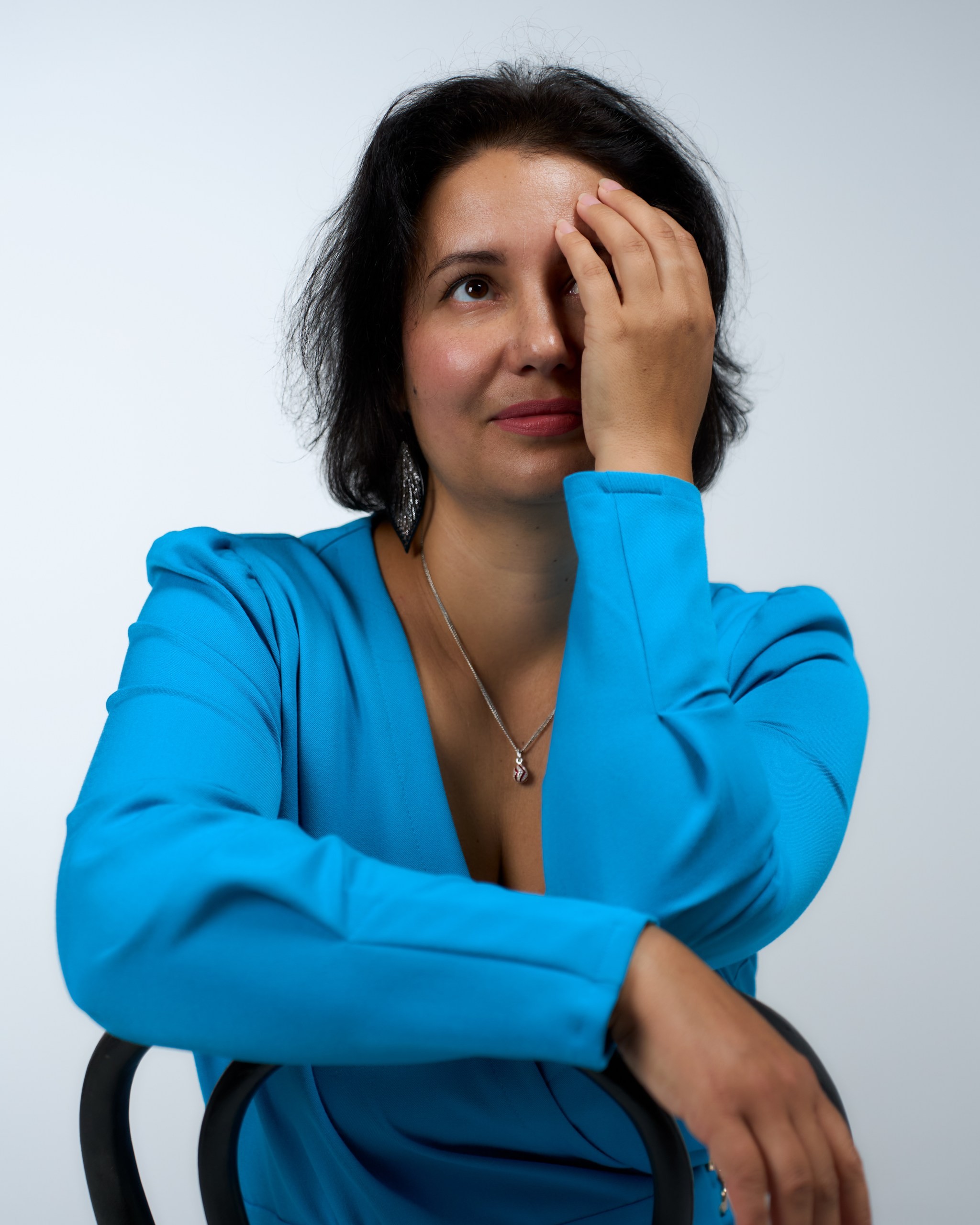 A person in a blue dress sits in a chair, winking with one eye covered by their hand, looking upward.