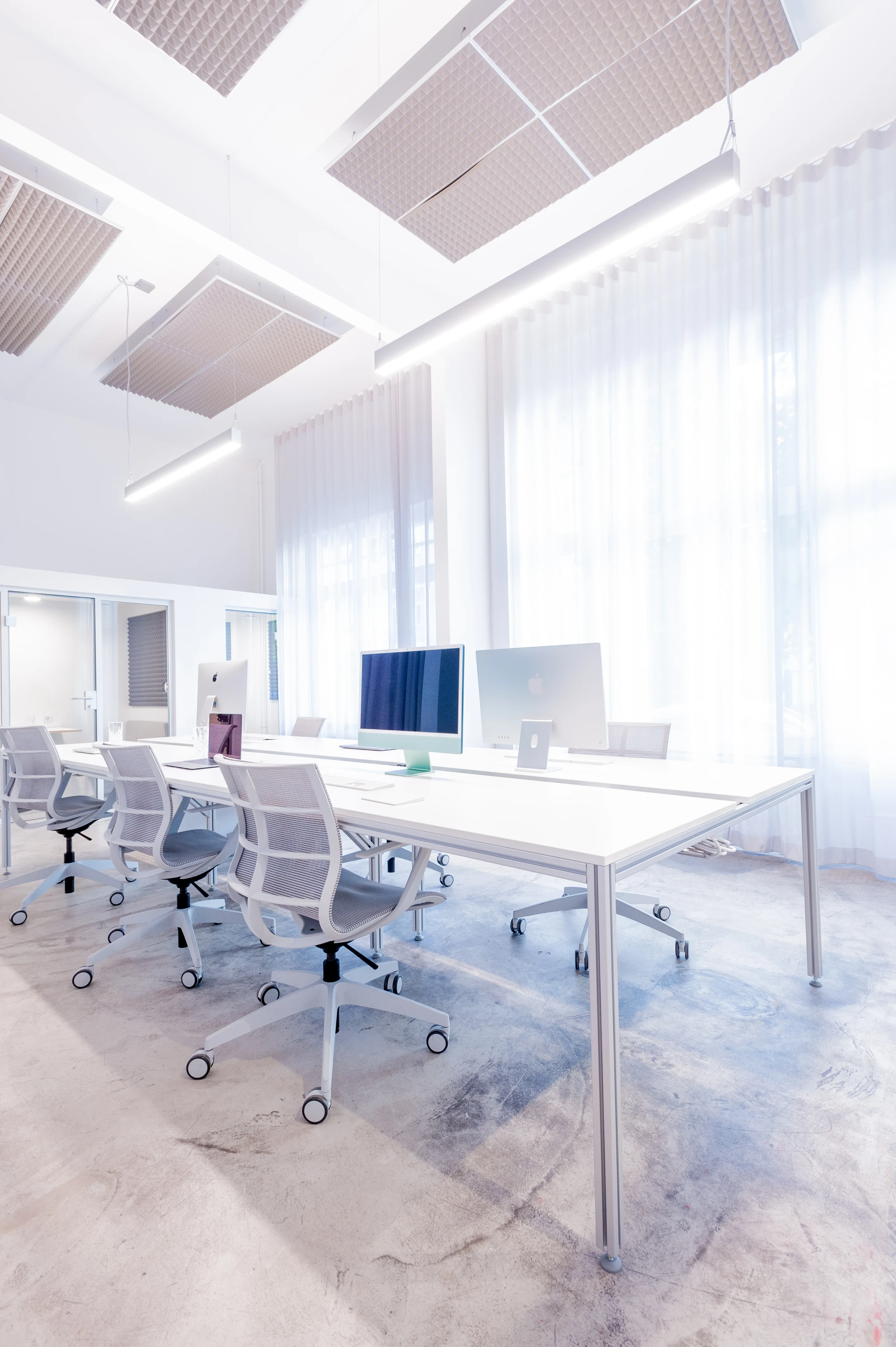 Bright all-white office at BATTLE ROYAL with white desks and mesh task chairs, iMac screens, sheer floor-length curtains, polished concrete floor and suspended perforated acoustic ceiling panels.