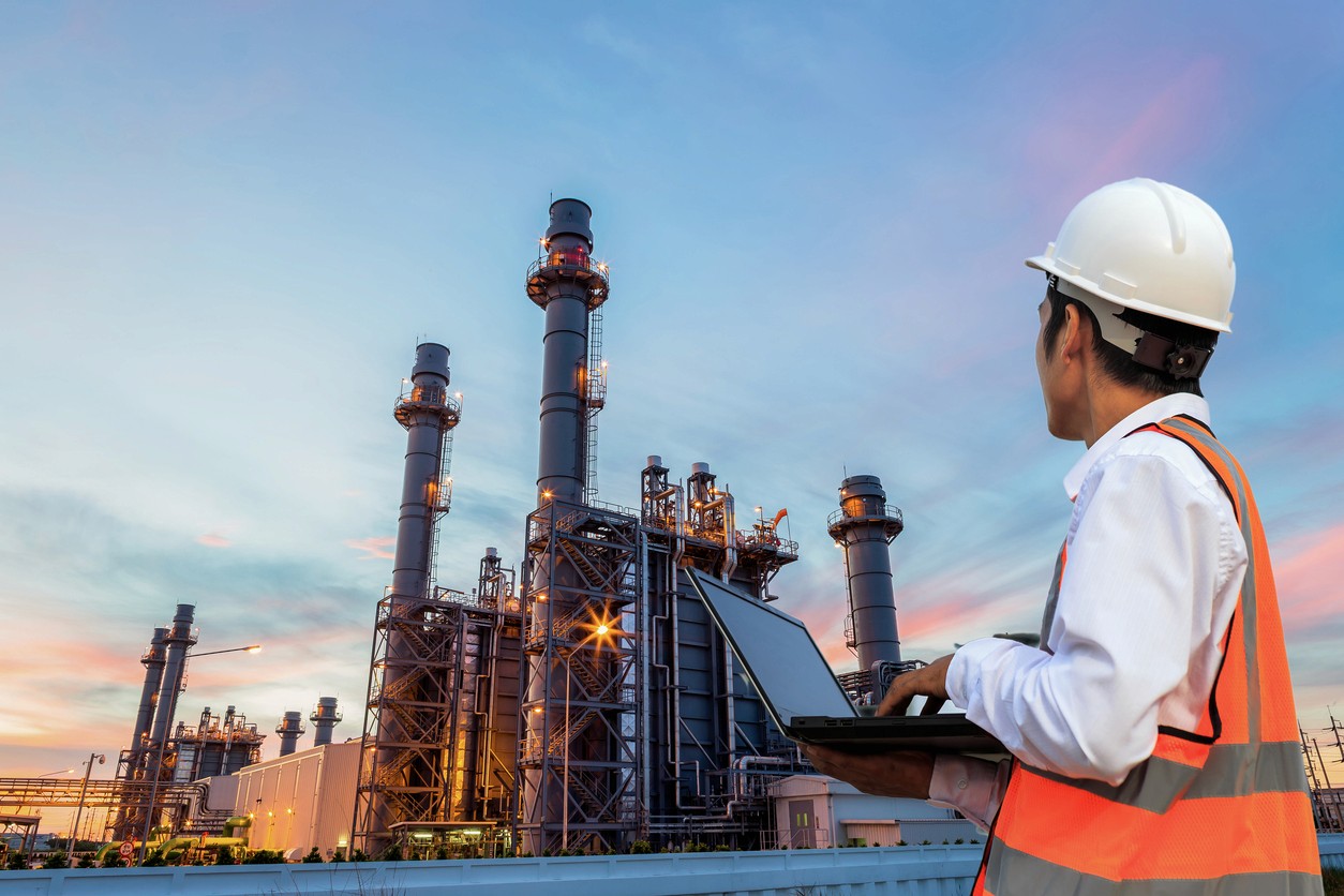 A construction worker in a hard hat observes an industrial facility at sunset, wearing safety gear.