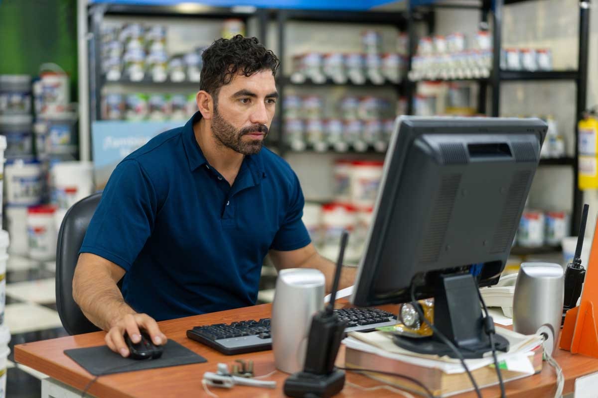 Comerciante usando um computador no balcão de uma loja de tintas e materiais de construção, com produtos organizados nas prateleiras ao fundo.