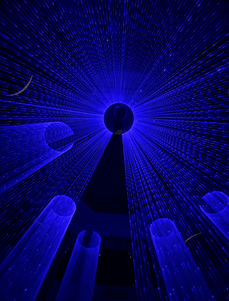 Instalação artística de luzes em tons de azul intenso, com feixes convergindo para uma esfera central iluminada.