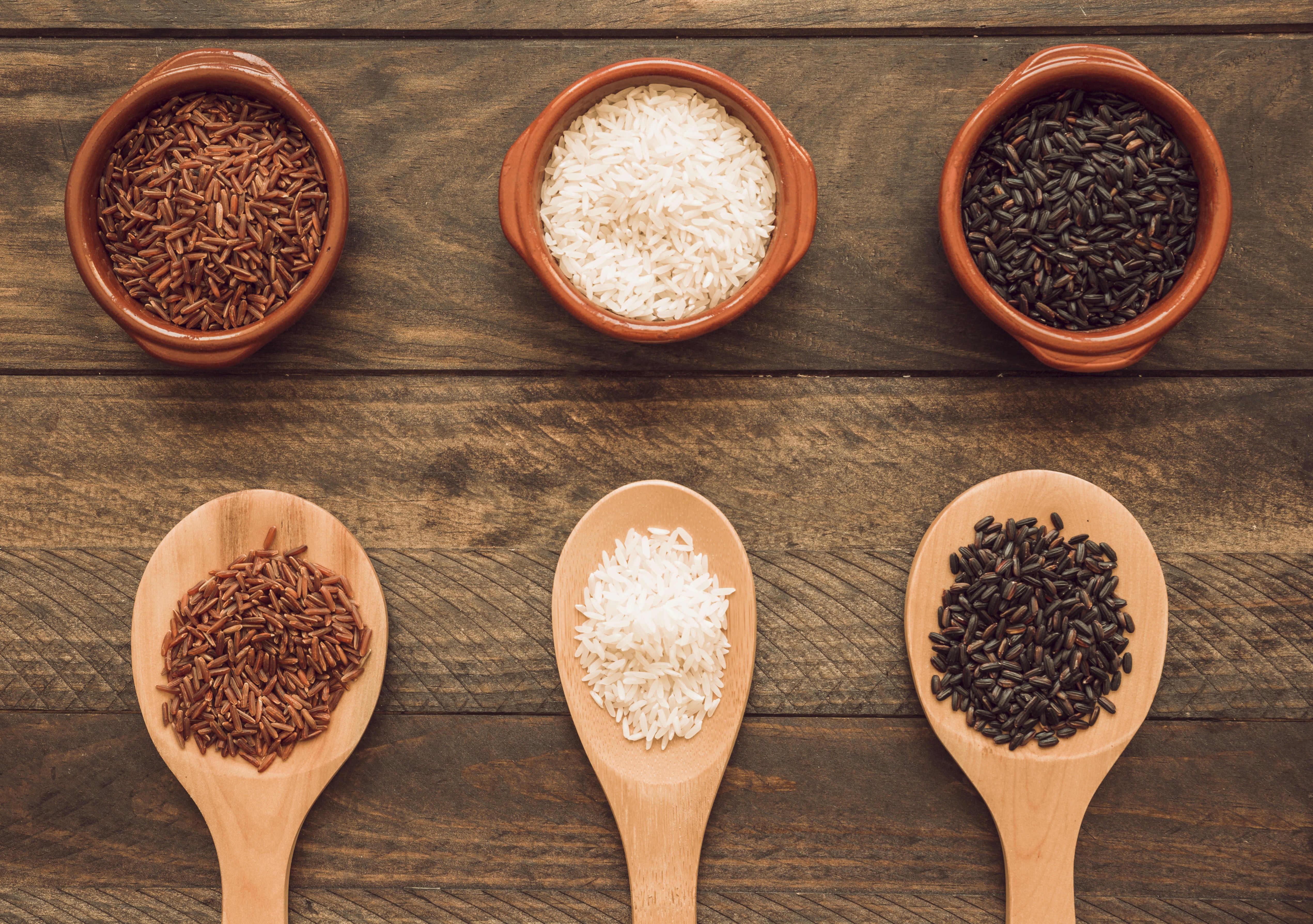 Three small bowls and wooden spoons display red, white, and black rice on a wooden table, evoking a rustic, earthy ambiance.