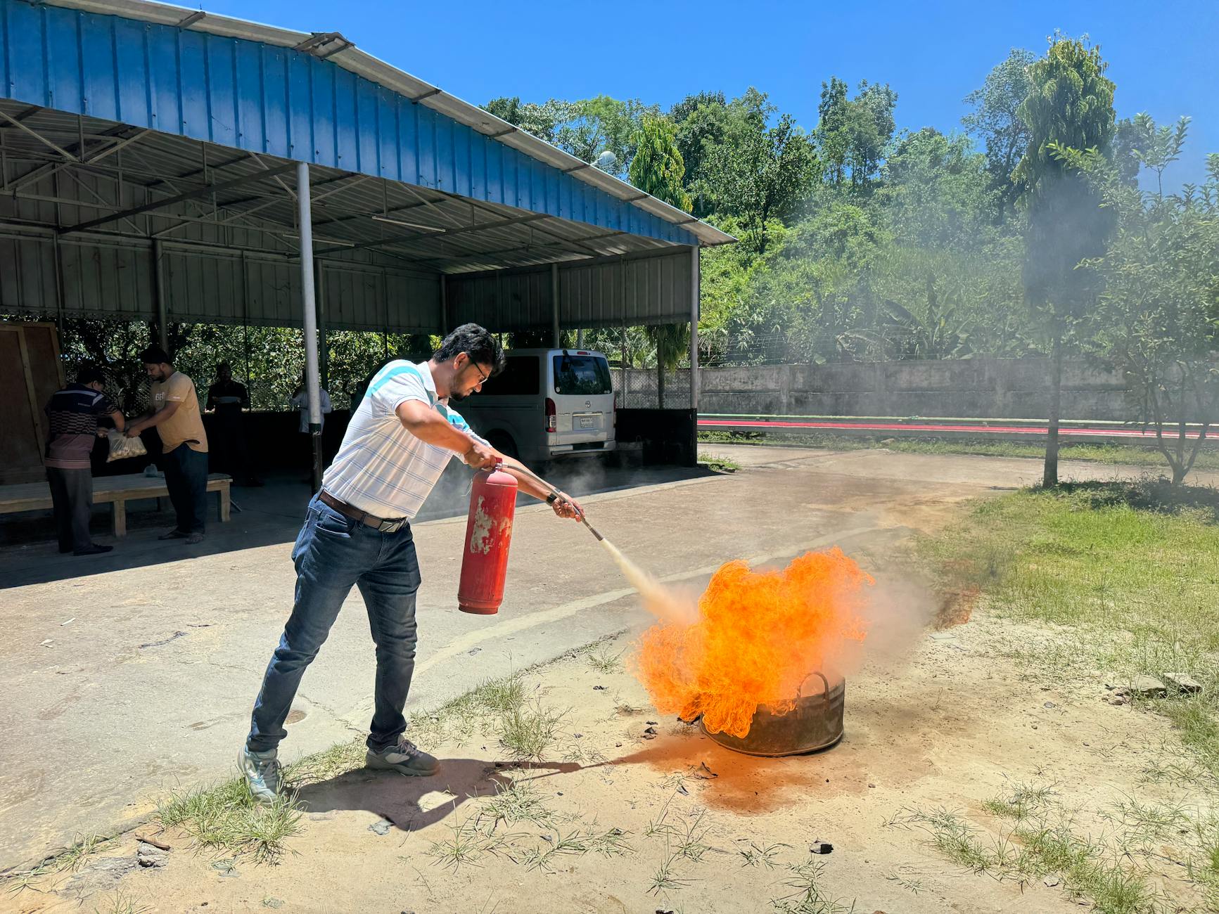 Man in white t-shirt using a red fire extinguisher on a ground-level fire in an outdoor concrete yard