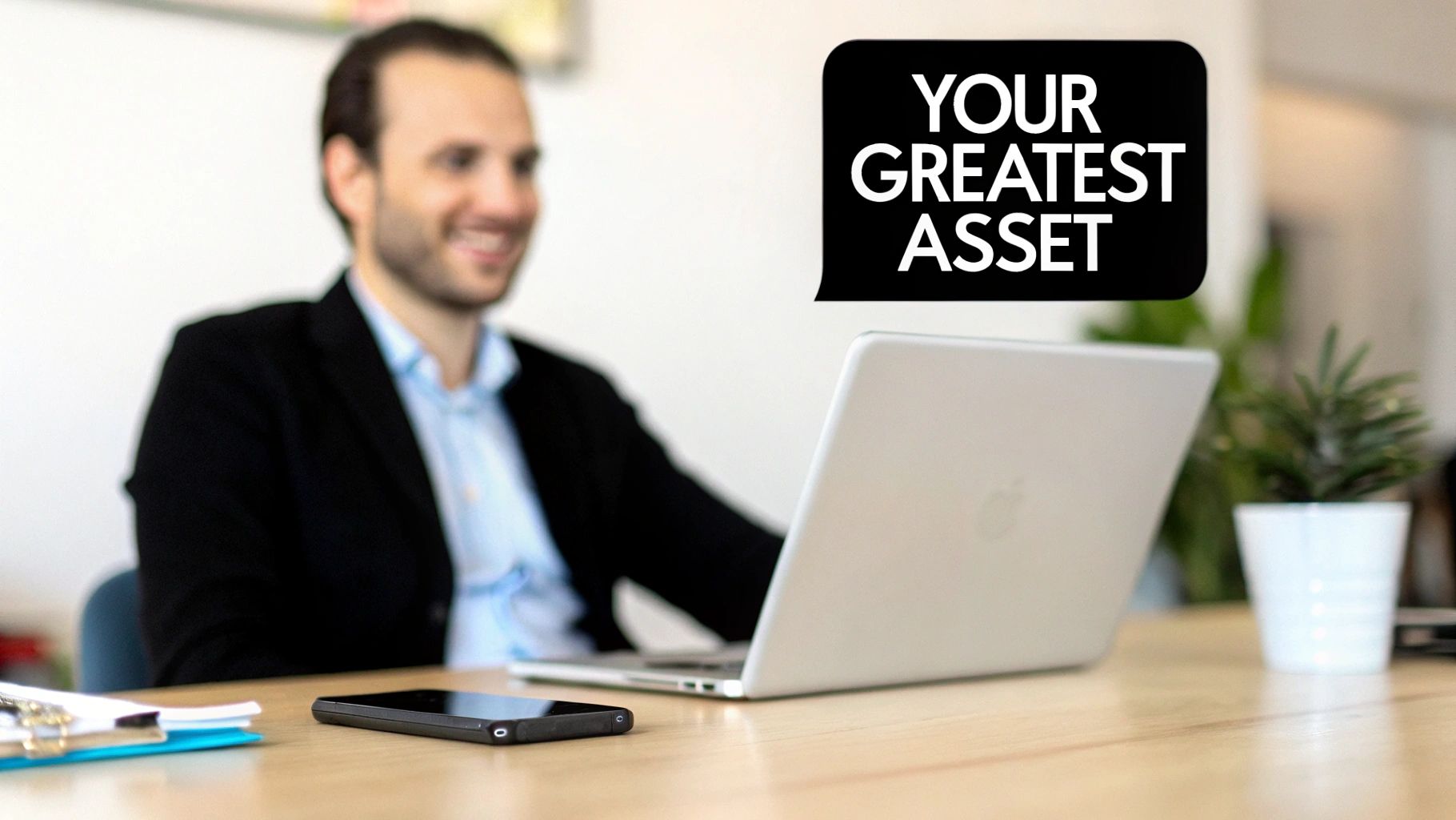Smiling businessman at a desk with a laptop and smartphone, speech bubble says 'YOUR GREATEST ASSET'.