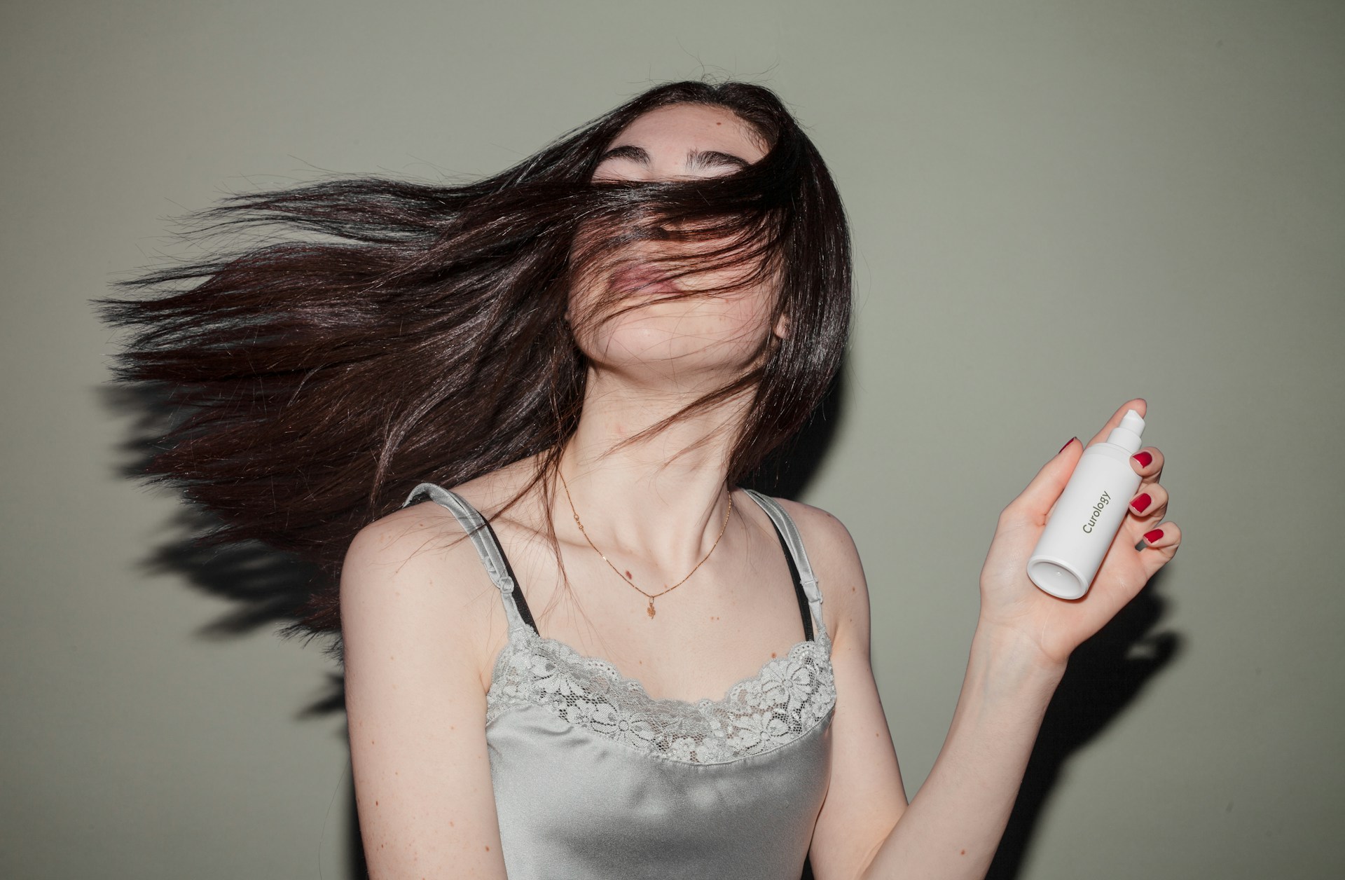 A young woman with long dark hair holds a white spray bottle and turns her head, sending her hair flying around her face.