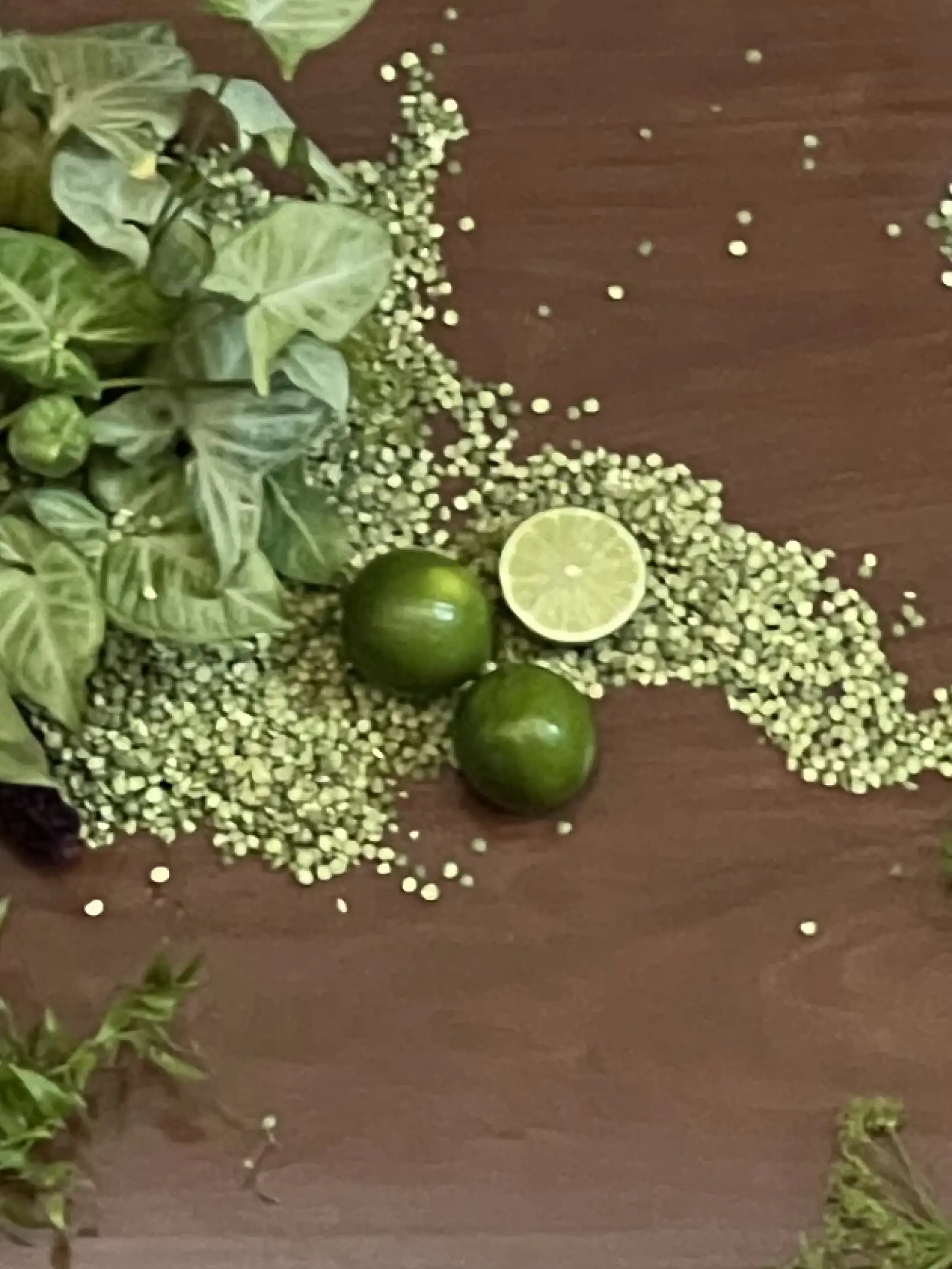 Detalle de limas y hojas verdes sobre mesa de madera con textura orgánica y granos esparcidos como parte de la ambientación natural.