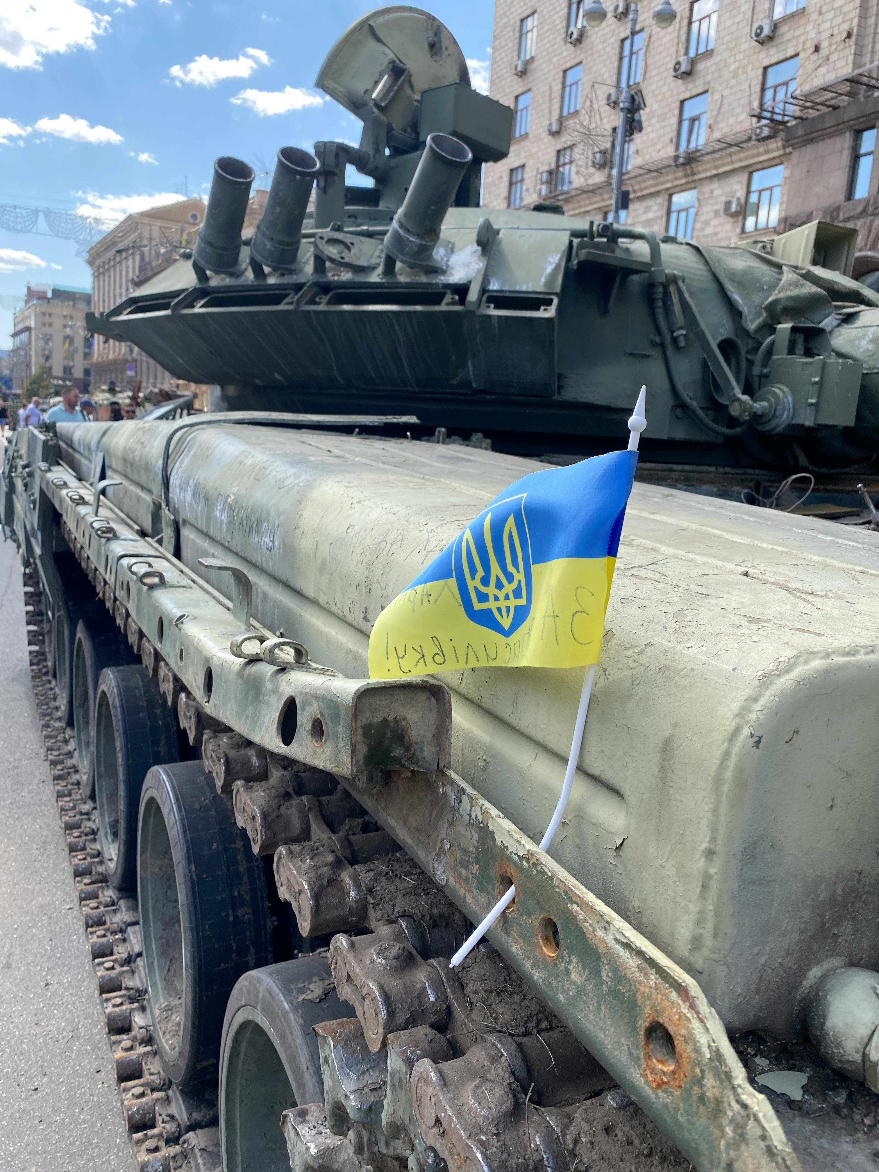 A military tank or armored vehicle on display in an urban setting with a small Ukrainian flag mounted on it. The heavy military equipment appears to be on public display, with buildings visible in the background. The blue and yellow Ukrainian flag with the trident emblem is prominently featured.