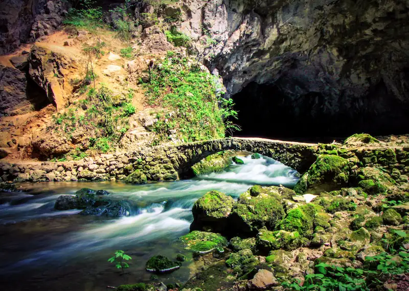 A small stream flowing under a stone bridge with a cave entrance in the background at Rakov Škocjan, Slovenia