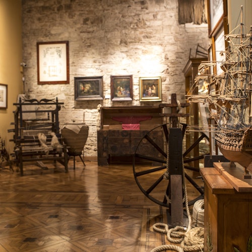 Museum room with wooden floor, ship models, vintage furniture, maritime artifacts, and framed paintings on a stone wall.
