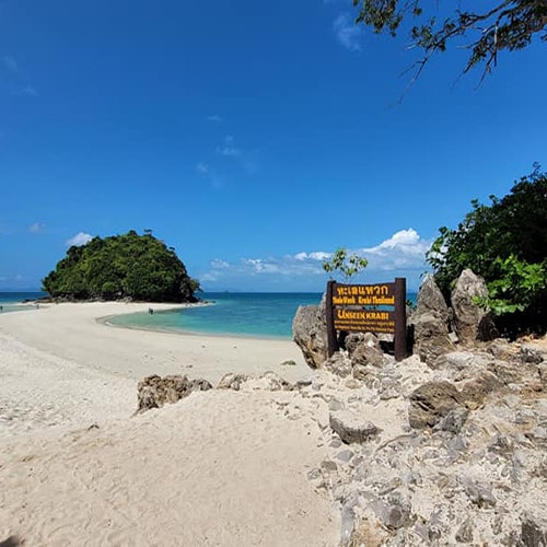 阳光明媚的海滩，海水清澈，远处有一个小岛。在岩石中间立着一块标有泰文和英文的指示牌。