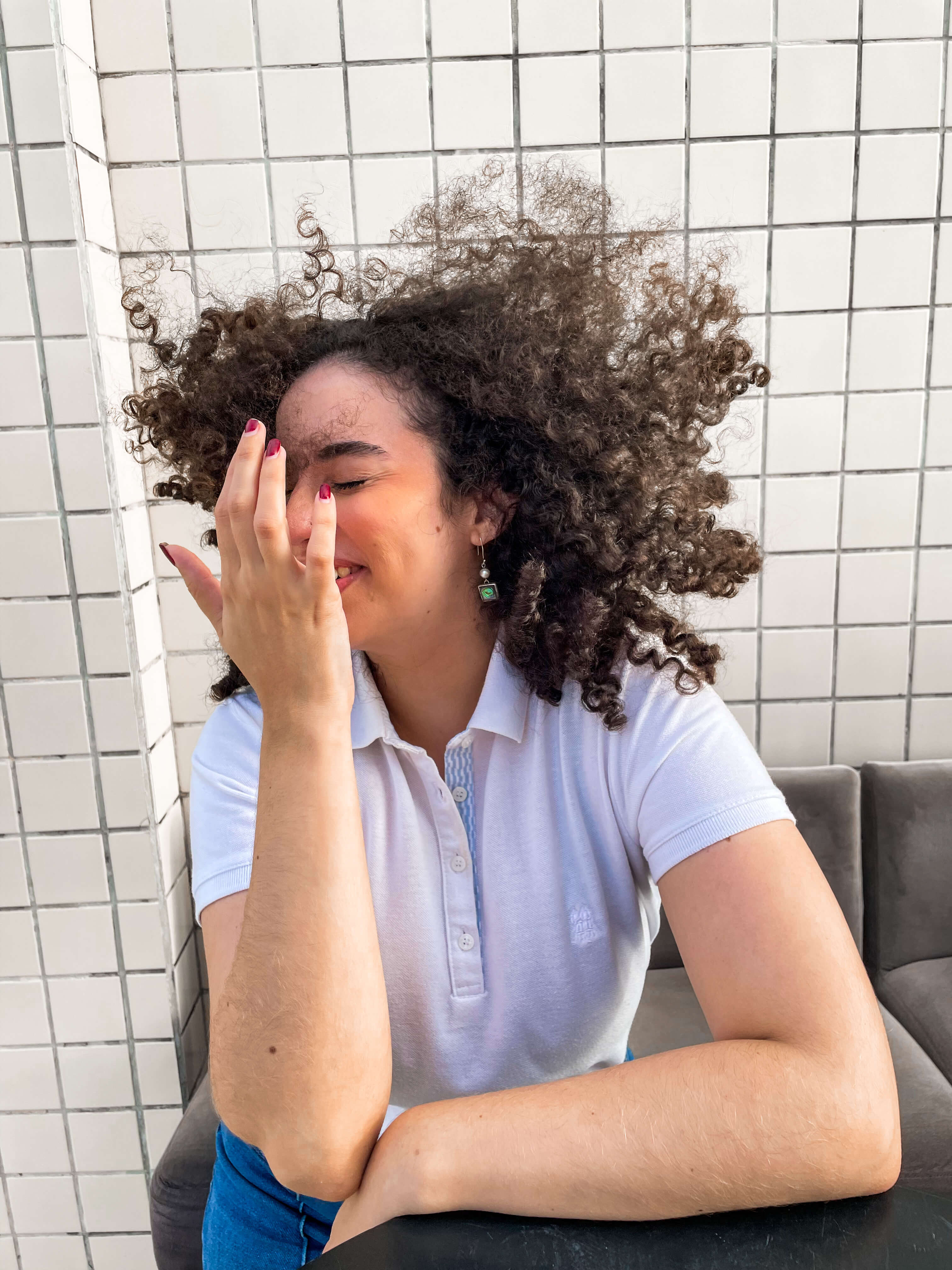 Young woman with curly dark hair blown up around her face while she smiles, touching her finger to her forehead